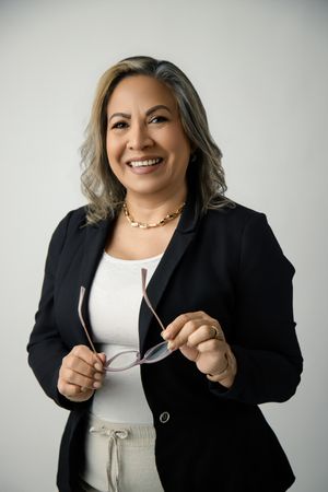 Smiling woman with wavy highlighted hair, wearing a black blazer and gold necklace, holds her glasses against a plain light background.