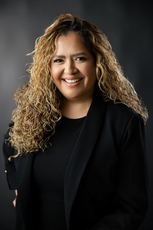 Smiling woman with long, curly highlighted hair wearing a black blazer over a black top against a dark gray studio background.