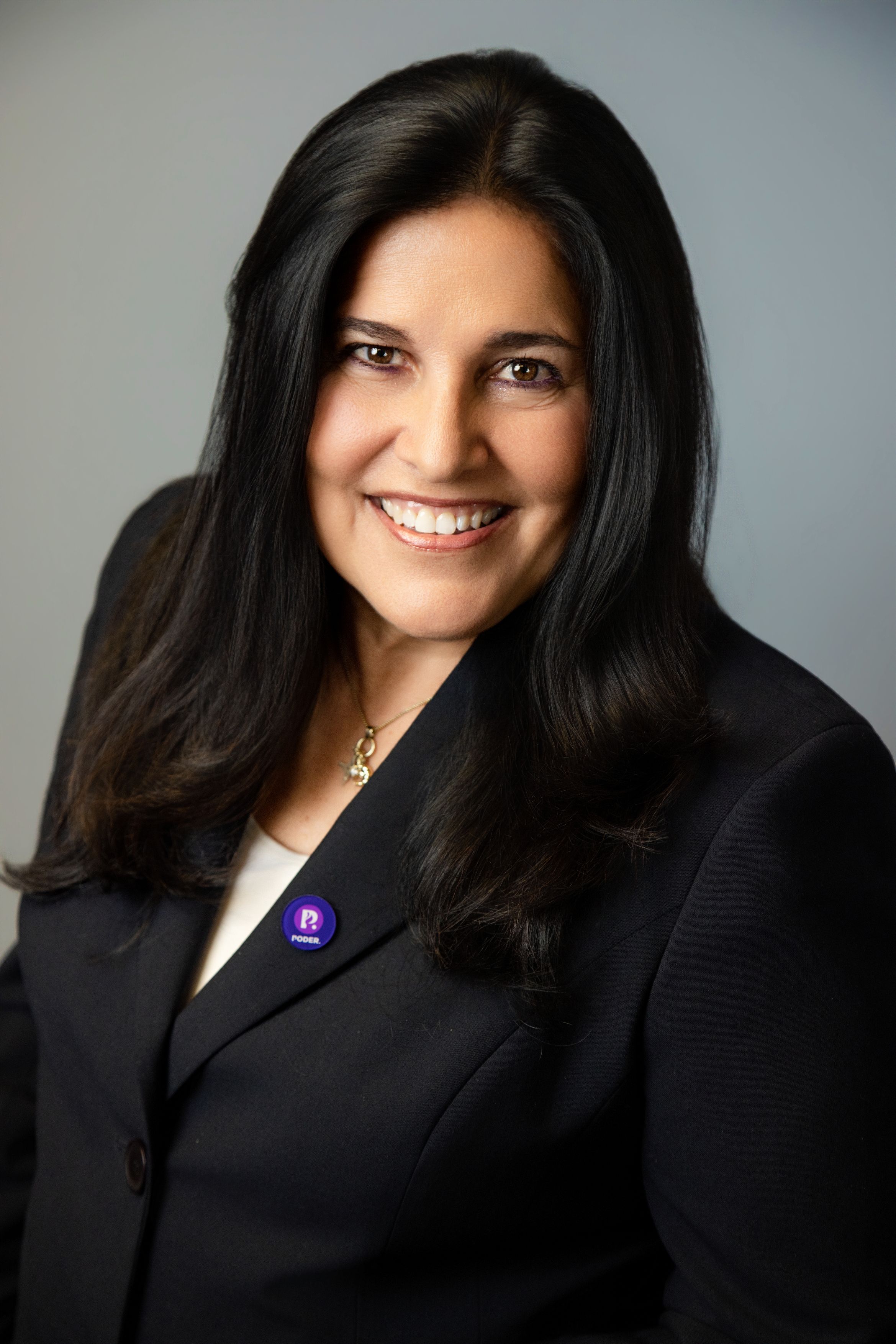 Smiling woman with long dark hair wearing a black blazer, a pendant necklace and a small purple pin in a professional headshot.