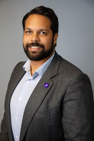 Smiling bearded man in a gray blazer and light blue collared shirt with a small purple pin on his lapel, posed against a gray backdrop.