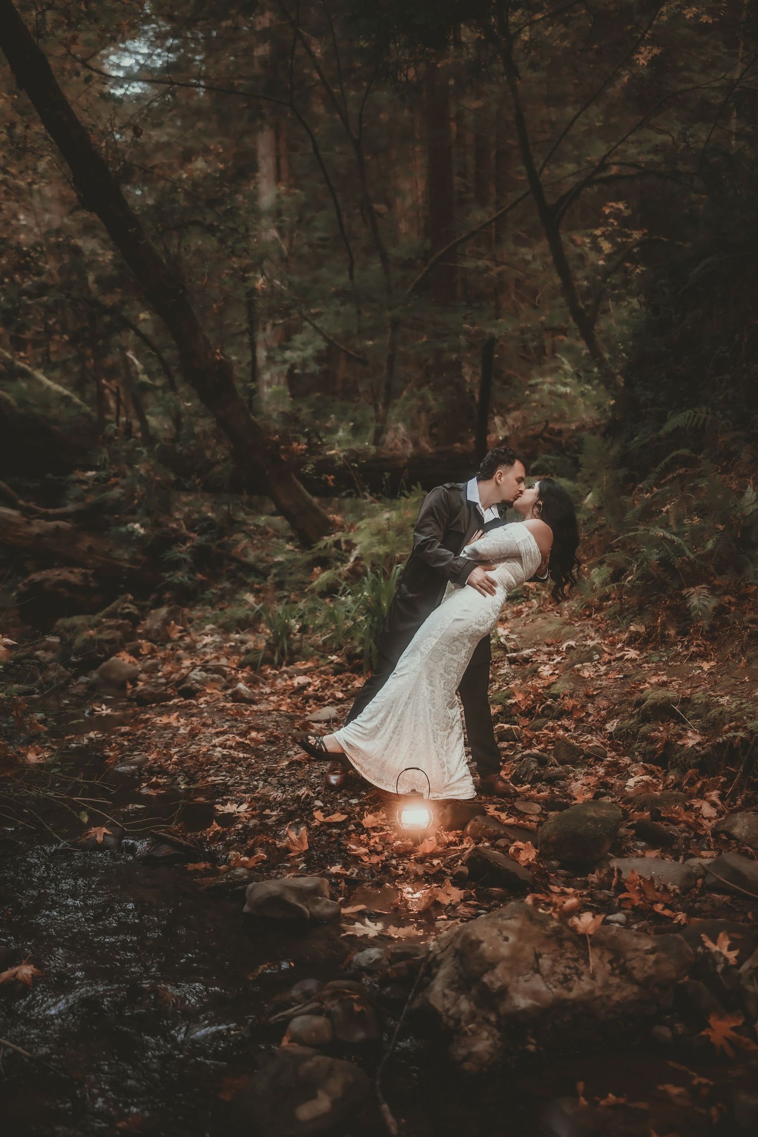 Couple in wedding attire share a dramatic dip kiss in a moody redwood forest, with a glowing lantern and fallen autumn leaves at their feet.
