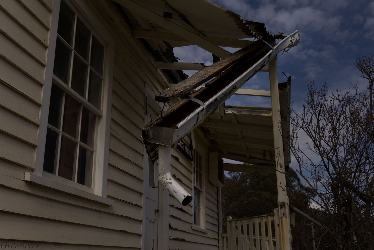 Weathered white wooden house with a collapsing porch roof, broken beams and peeling siding under a dramatic cloudy sky.