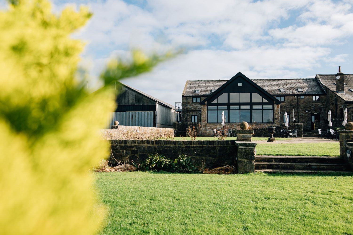 Stone venue building with large gabled glass windows overlooking a neatly mown green lawn, with blurred yellow foliage in the foreground.