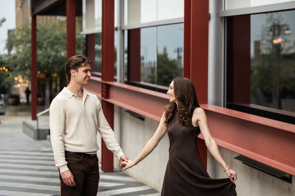 Smiling couple holding hands outdoors beside a modern red-trimmed glass building, the man in a cream sweater and the woman in a brown dress.