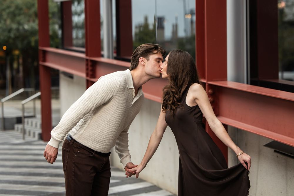 Romantic couple sharing a kiss while holding hands outdoors beside a modern building with red steel beams and glass windows.