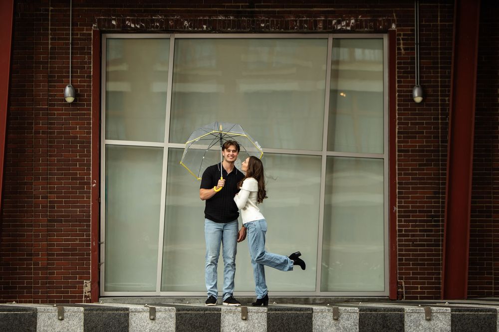 Young couple kissing under a clear umbrella, the woman lifting her leg playfully, in front of a large window and brick building.