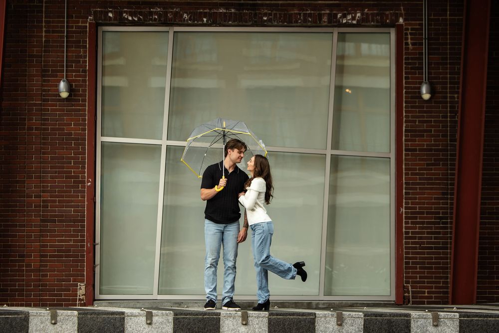 Couple sharing a romantic kiss under a clear umbrella in front of a large window and brick wall, with the woman lifting her foot.