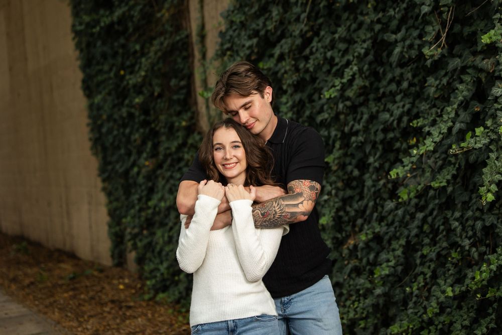 A smiling couple embraces in front of an ivy-covered wall, the man with tattoos hugging the woman in a white sweater from behind.