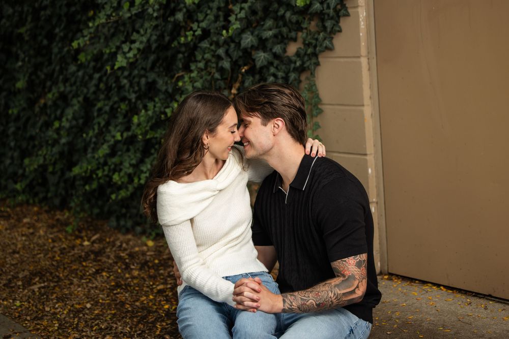 Couple sitting close together touching noses and holding hands in front of an ivy-covered wall during an outdoor engagement photo session.