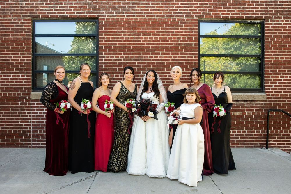 Bride in a white gown holding a bouquet poses with bridesmaids in burgundy, red, and black dresses and a flower girl before a brick wall.