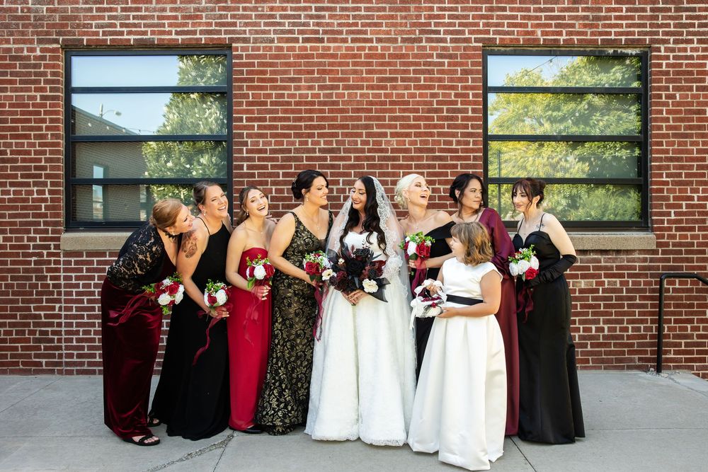 Bride in a white gown laughs with her bridesmaids in black, red, and burgundy dresses holding bouquets in front of a brick wall.