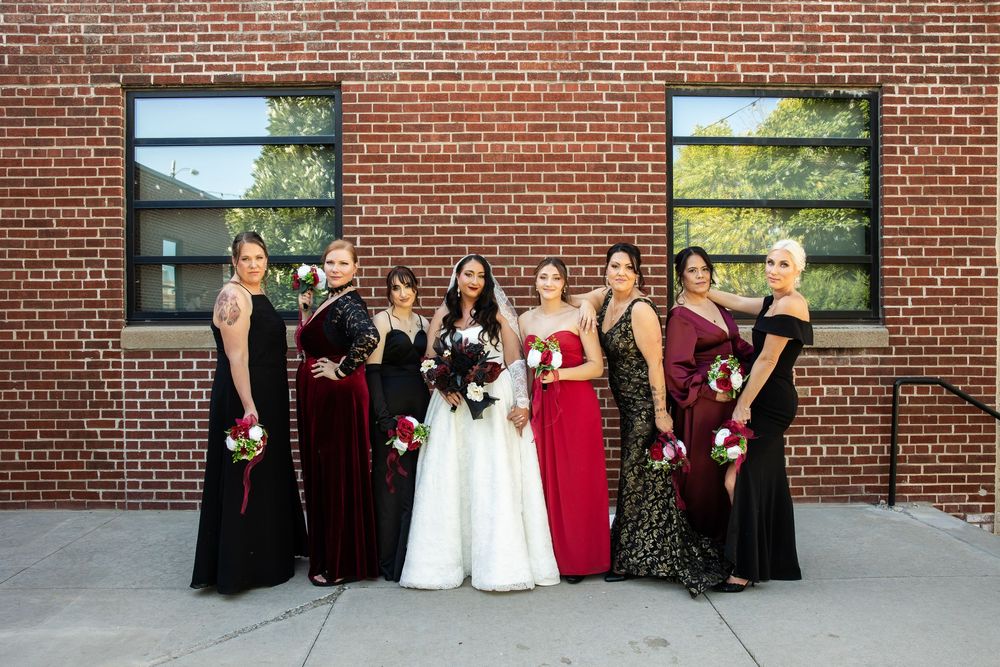 Bride in a white gown holding a bouquet stands among seven bridesmaids in black, burgundy and red dresses in front of a brick building.