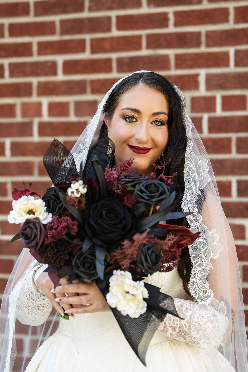 Bride in a lace-trimmed veil and white gown holding a gothic bouquet of black roses and burgundy blooms in front of a brick wall.