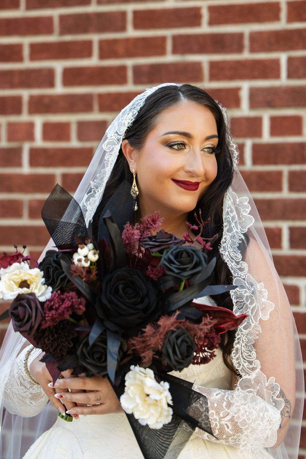 Smiling bride in a lace-trimmed veil holds a moody bouquet of black roses, burgundy blooms and a white flower against a red brick wall.