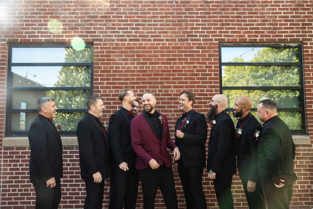 Groomsmen in dark suits, one in a burgundy jacket, laugh together while standing against a red brick wall at a wedding.