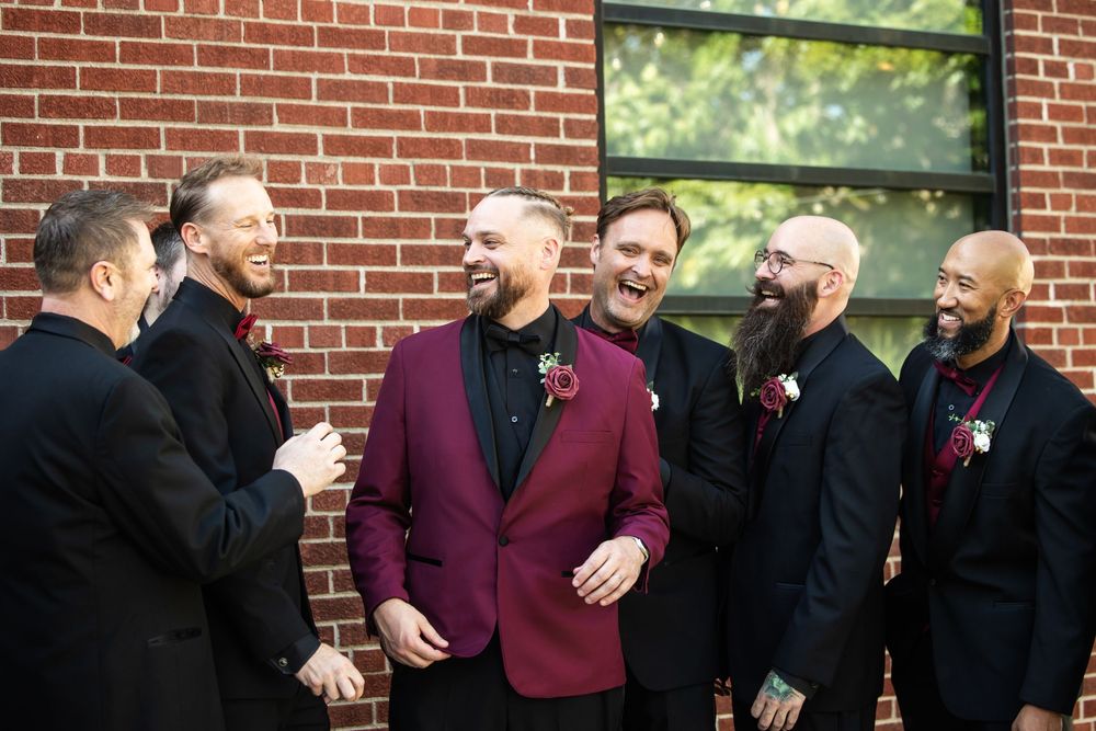 Six laughing groomsmen in dark suits and floral boutonnieres, one in a burgundy velvet blazer, joking together against a red brick wall.