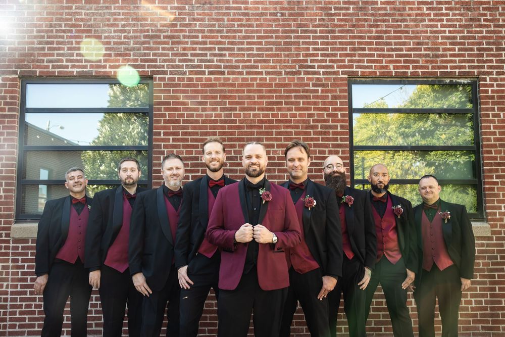 Nine groomsmen in black suits and burgundy vests with boutonnieres pose together in front of a red brick wall with large windows.