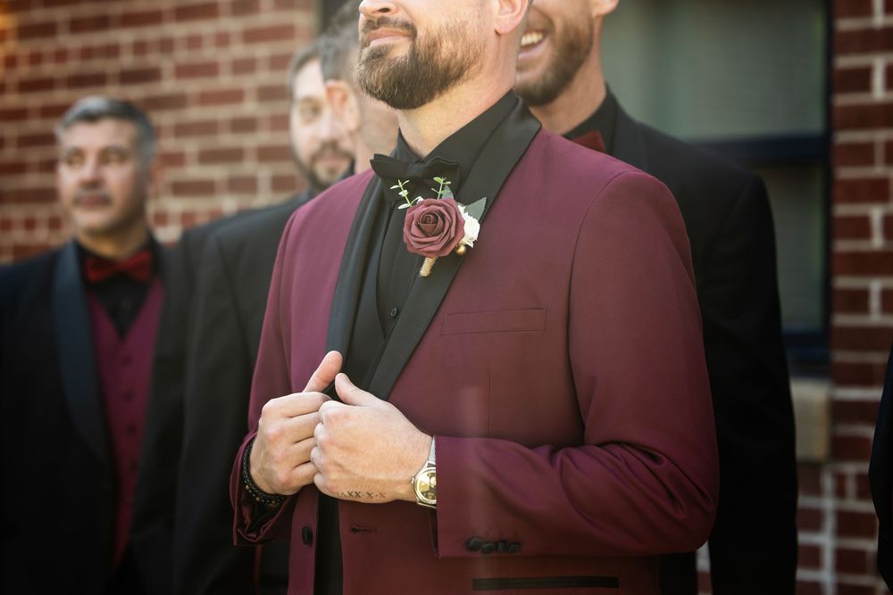 Groom in a burgundy suit jacket with a rose boutonniere adjusts his cuff while groomsmen in black stand behind him near a brick wall.