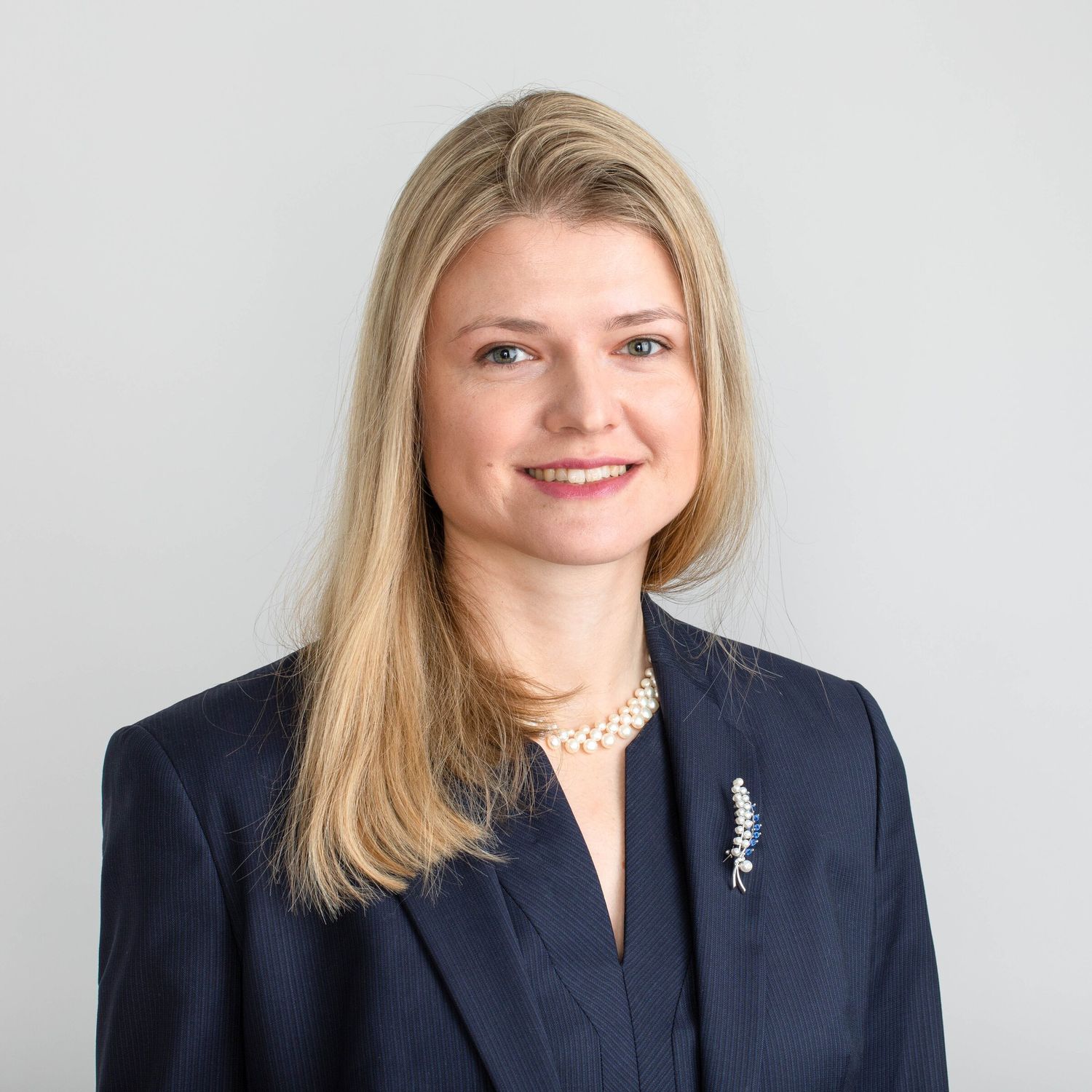 Professional headshot of a businesswoman in a navy jacket and pearl necklace on a light grey background