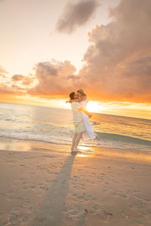 Couple sharing a romantic kiss on a beach at sunset, man lifting woman in white dress with golden sky backdrop.