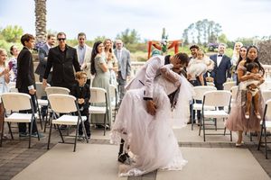 Groom dips bride in a romantic kiss at outdoor wedding ceremony while guests celebrate in the background.