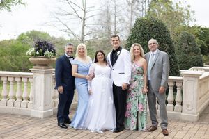 Wedding party of six poses on elegant stone terrace with balustrade, bride in white gown, guests in formal attire.