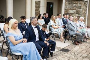Guests seated in chairs at an outdoor wedding ceremony, dressed in formal attire including blue dresses and suits.