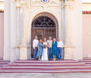 Wedding party of six posing on church steps in front of ornate arched doors, bride in white gown with bouquet.
