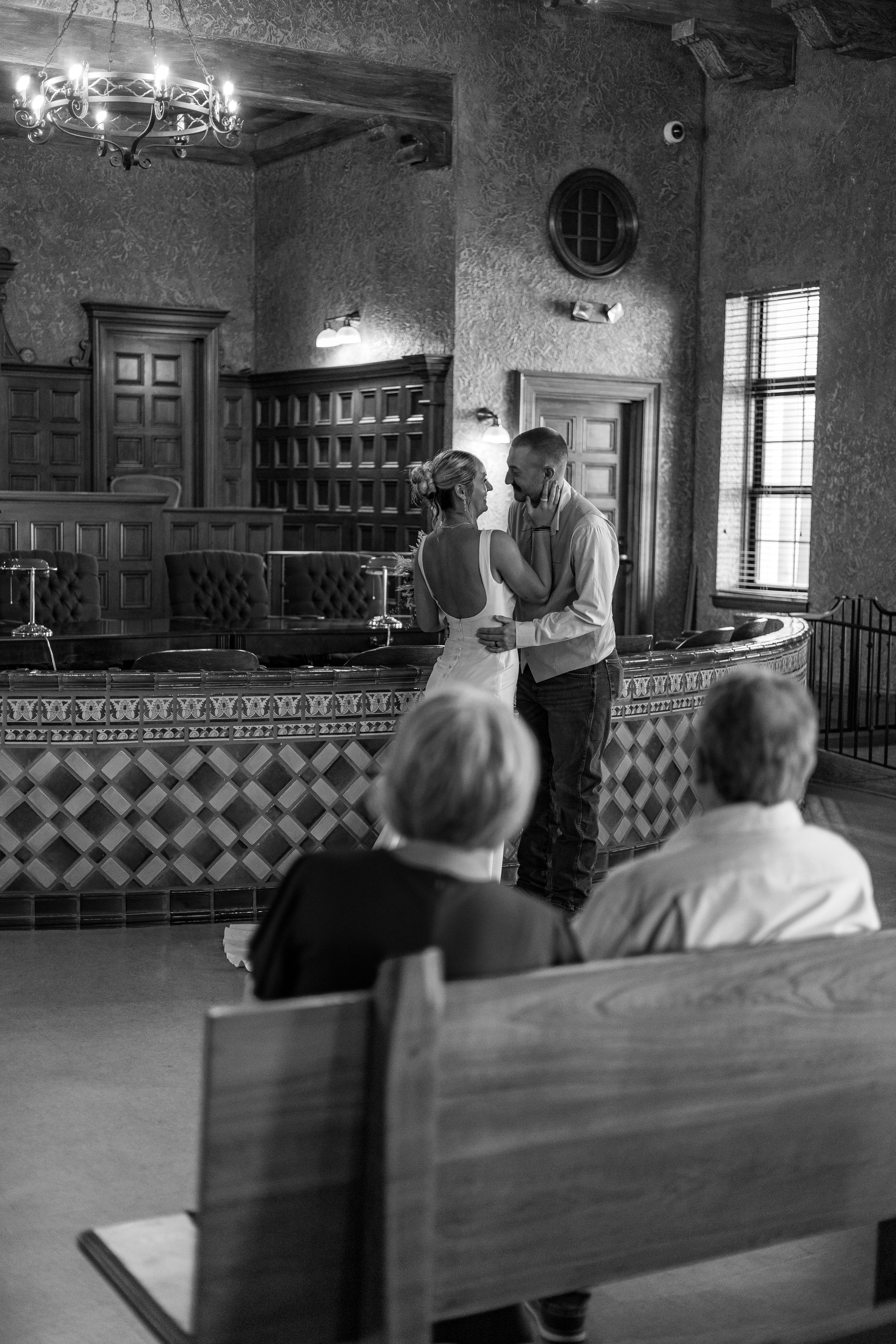 Black and white photo of a couple sharing an intimate moment during a small courthouse wedding ceremony with guests seated nearby.