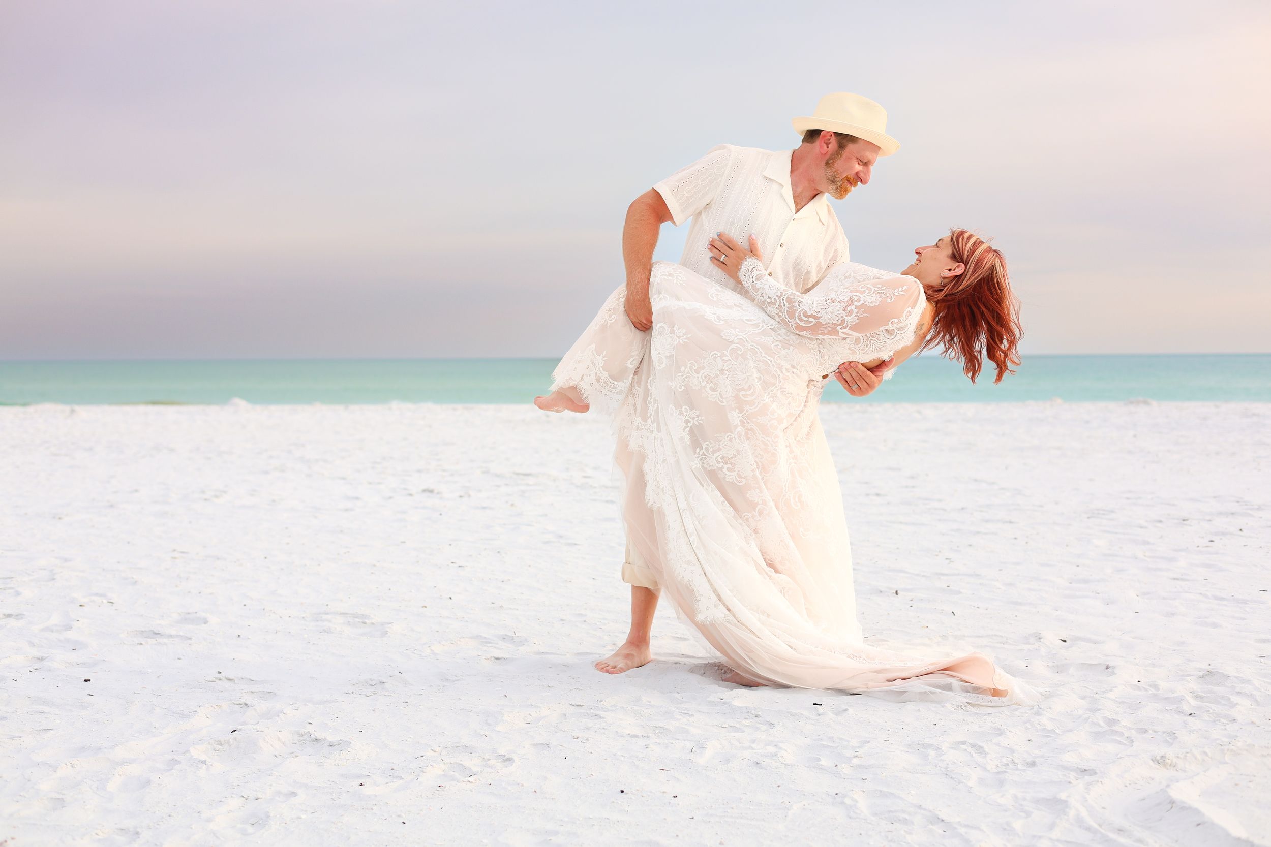 Couple in white attire sharing a romantic dip dance pose on a serene white sand beach at sunset.