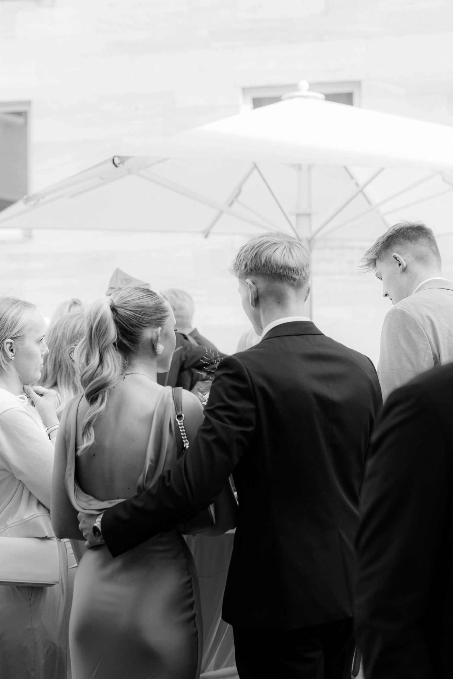 Black and white photo of elegantly dressed wedding guests gathered outdoors under a large patio umbrella.