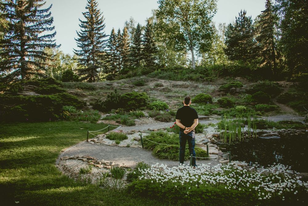 Man standing alone by a serene garden pond surrounded by lush greenery and tall evergreen trees.