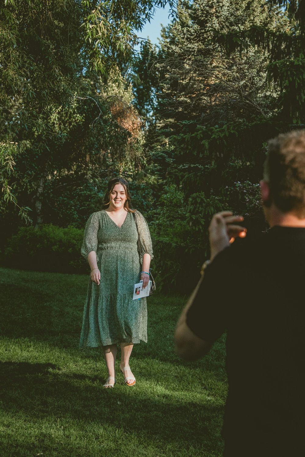 Woman in green dress smiling as man photographs her in a lush outdoor garden setting.