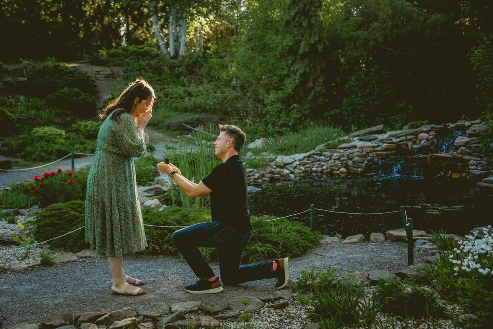 Man proposing on one knee to woman in green dress in a beautiful garden landscape.