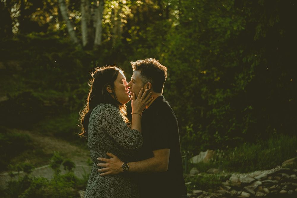 A couple shares a romantic kiss outdoors surrounded by lush green trees in warm golden light.