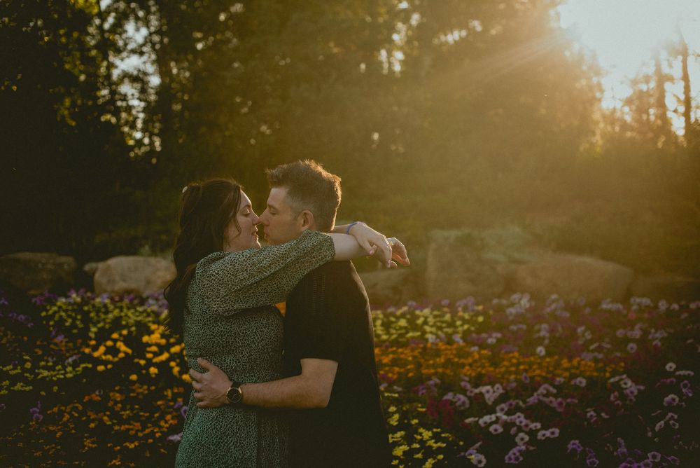 Couple embracing and kissing in a sunlit garden with colorful wildflowers and golden hour light filtering through trees.