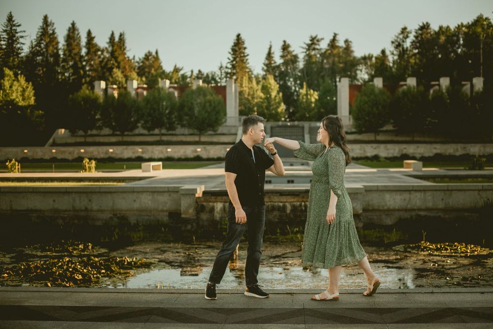 Couple dancing together near a reflective fountain in a sunlit park, woman in green dress and man in black outfit.