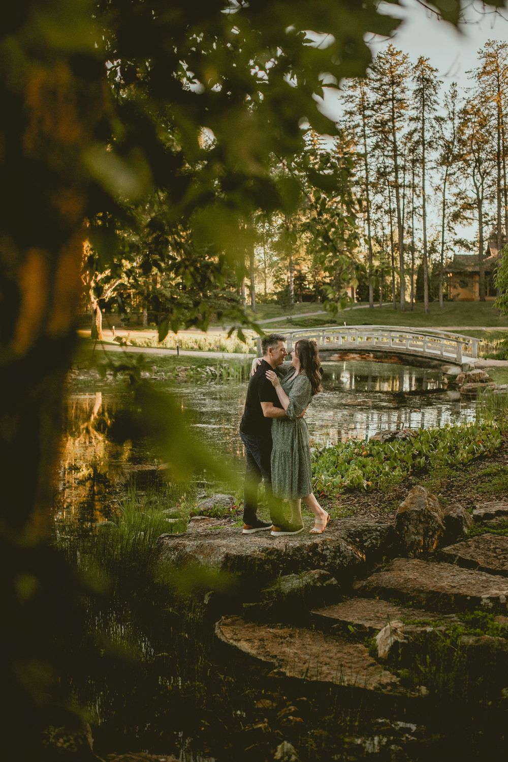 Couple embracing by a serene pond with a stone bridge, surrounded by lush greenery in golden hour light.