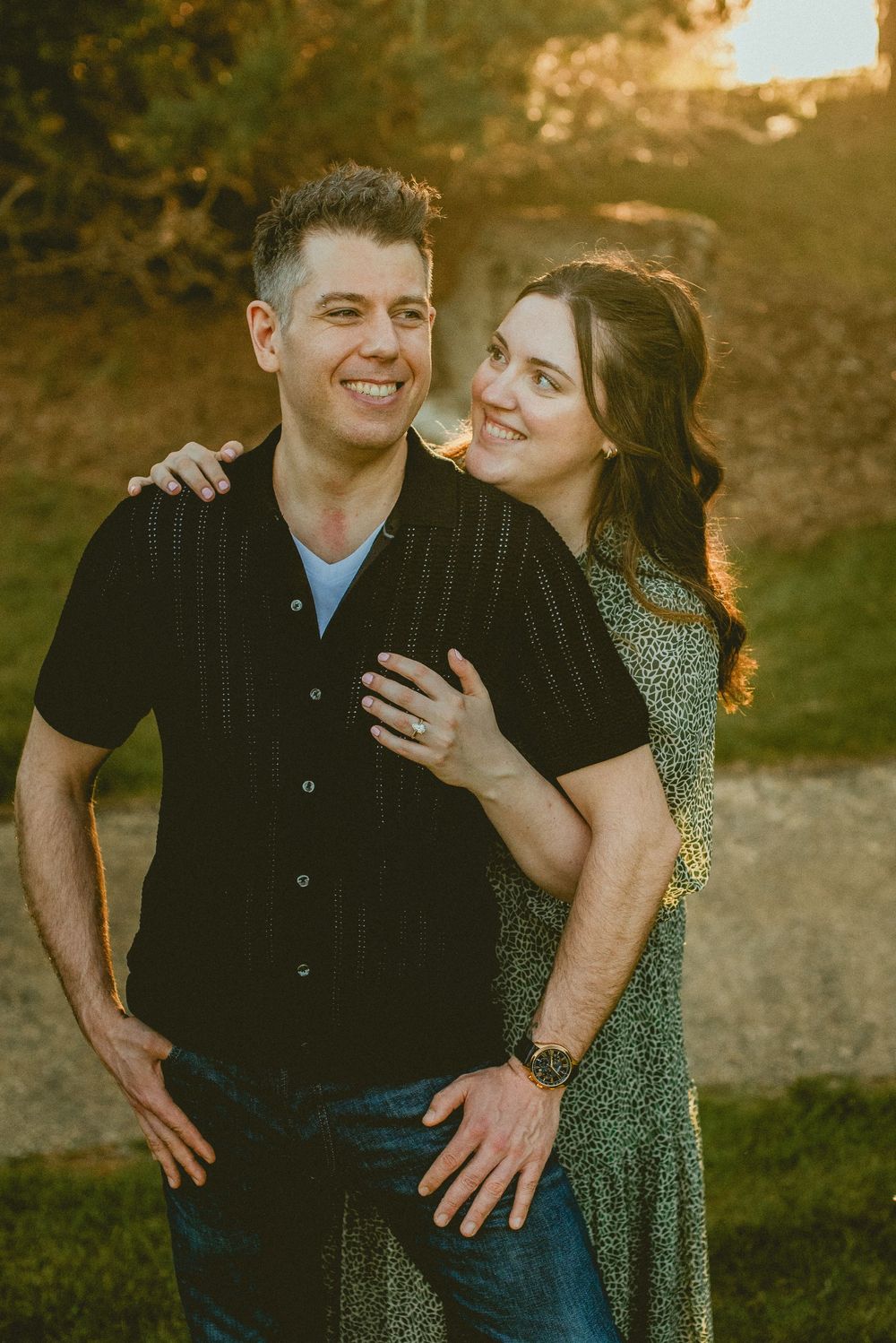 A smiling couple poses outdoors at golden hour, man in black shirt with woman in green floral dress embracing him.