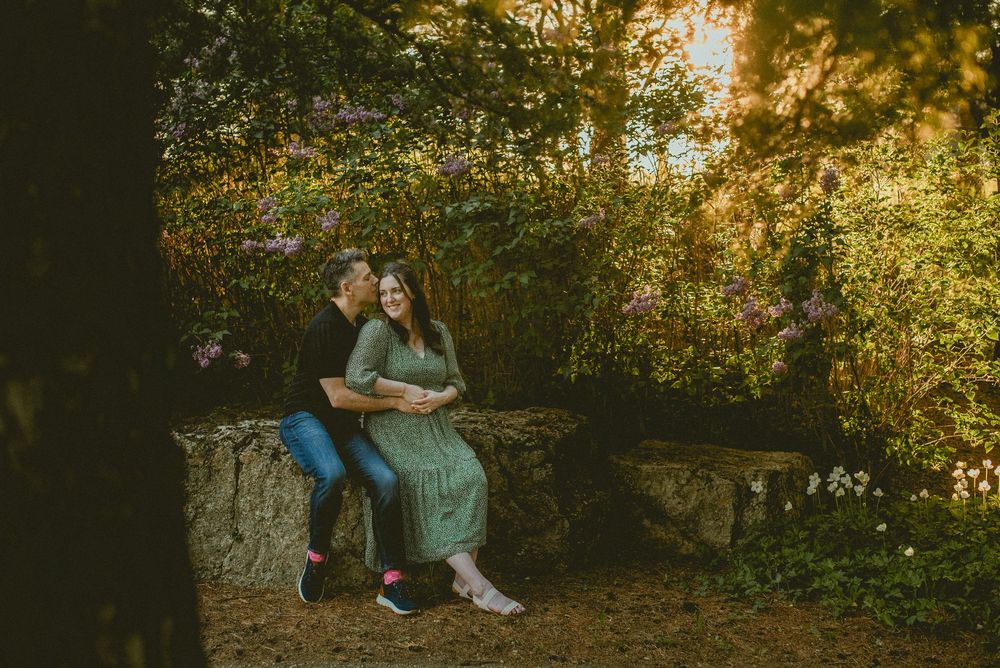Couple sitting on a stone bench surrounded by lush greenery and blooming flowers in a romantic outdoor garden setting.