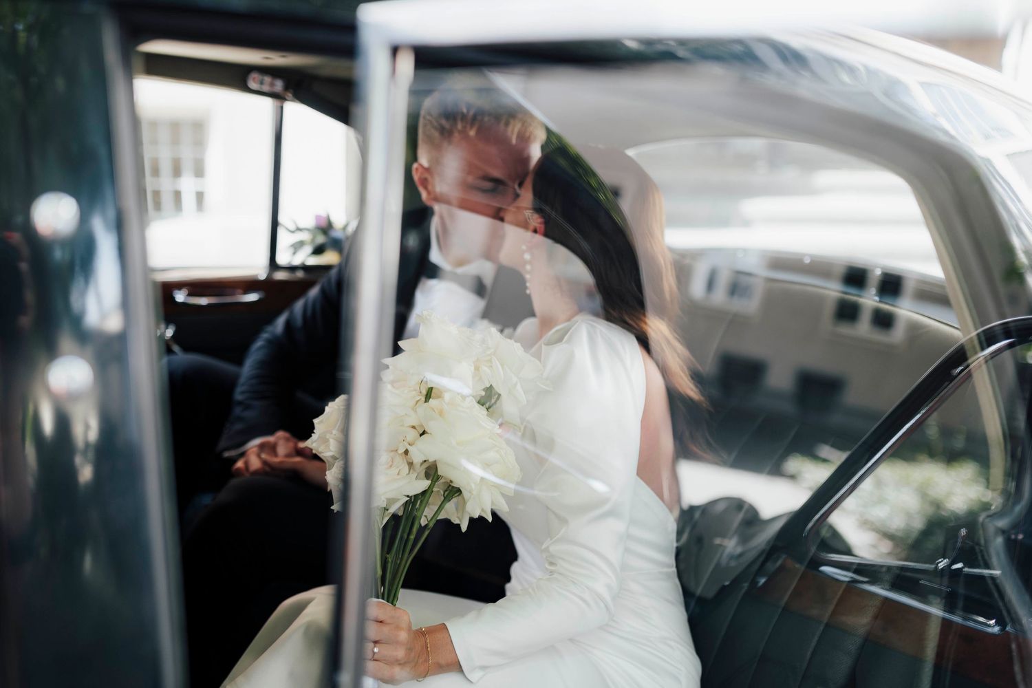 Newlywed couple kissing in a vintage black car, bride holding white bouquet in wedding dress.