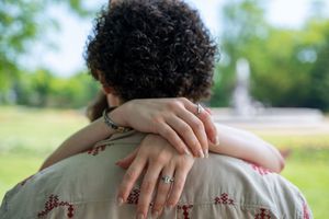 A woman with a ring on her finger embraces a curly-haired person from behind in a sunny park.