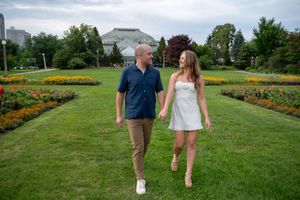 Couple holding hands walking through a colorful garden park with green lawn and flower beds on a cloudy day.