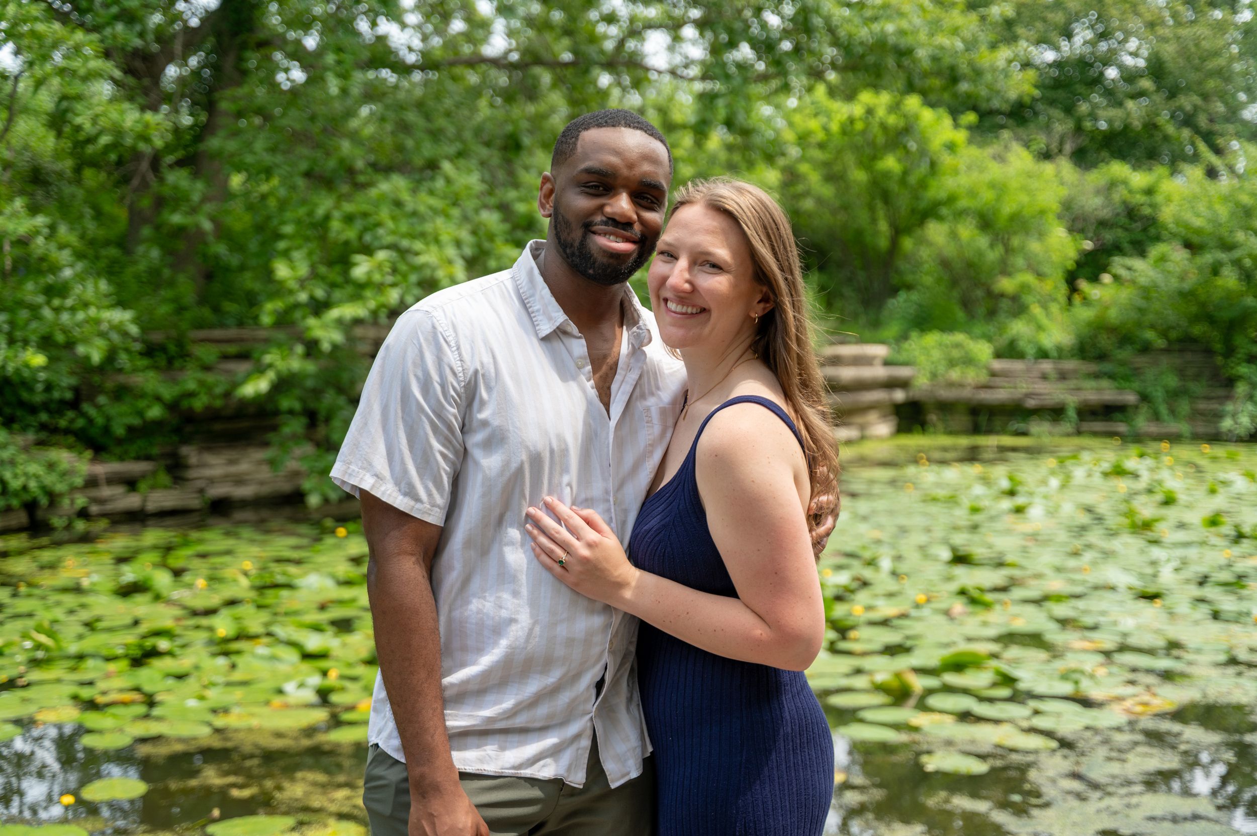 A smiling couple poses together in front of a serene lily pad pond surrounded by lush green trees.
