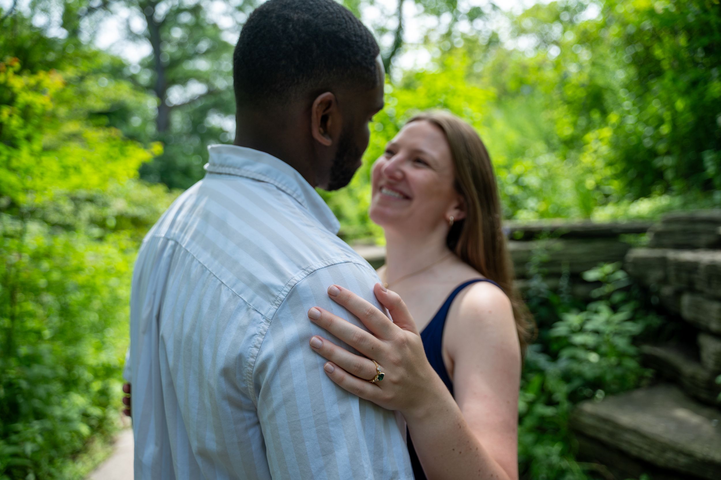 A smiling woman embraces a man outdoors, surrounded by lush green trees and foliage in a romantic engagement photo.