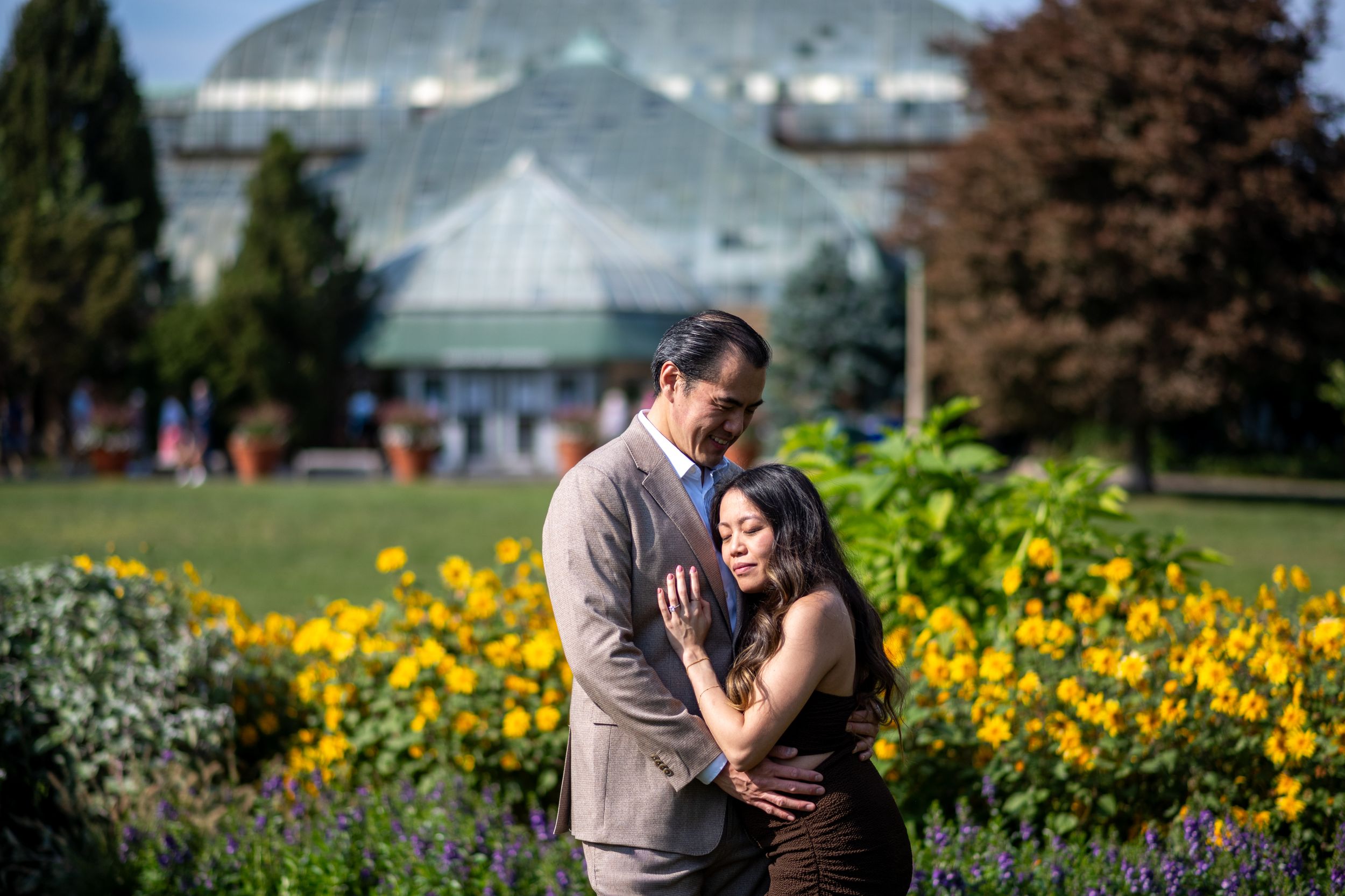 Couple embracing in a lush garden with yellow flowers and a glass greenhouse in the background on a sunny day.