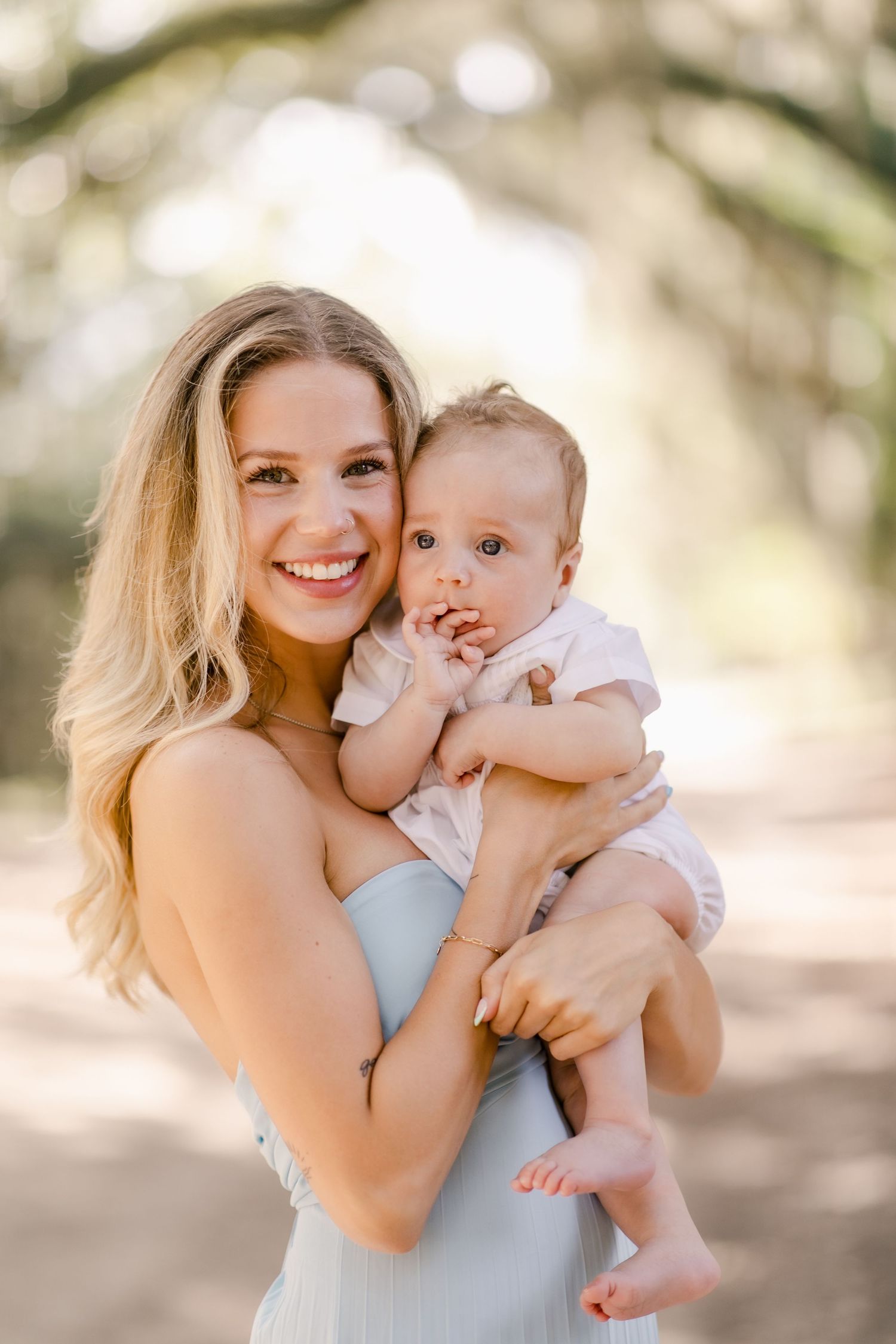 Smiling blonde woman holding a cute baby outdoors in a sun-dappled tree framed setting, with Spanish moss shining behind in St. Helena, SC.
