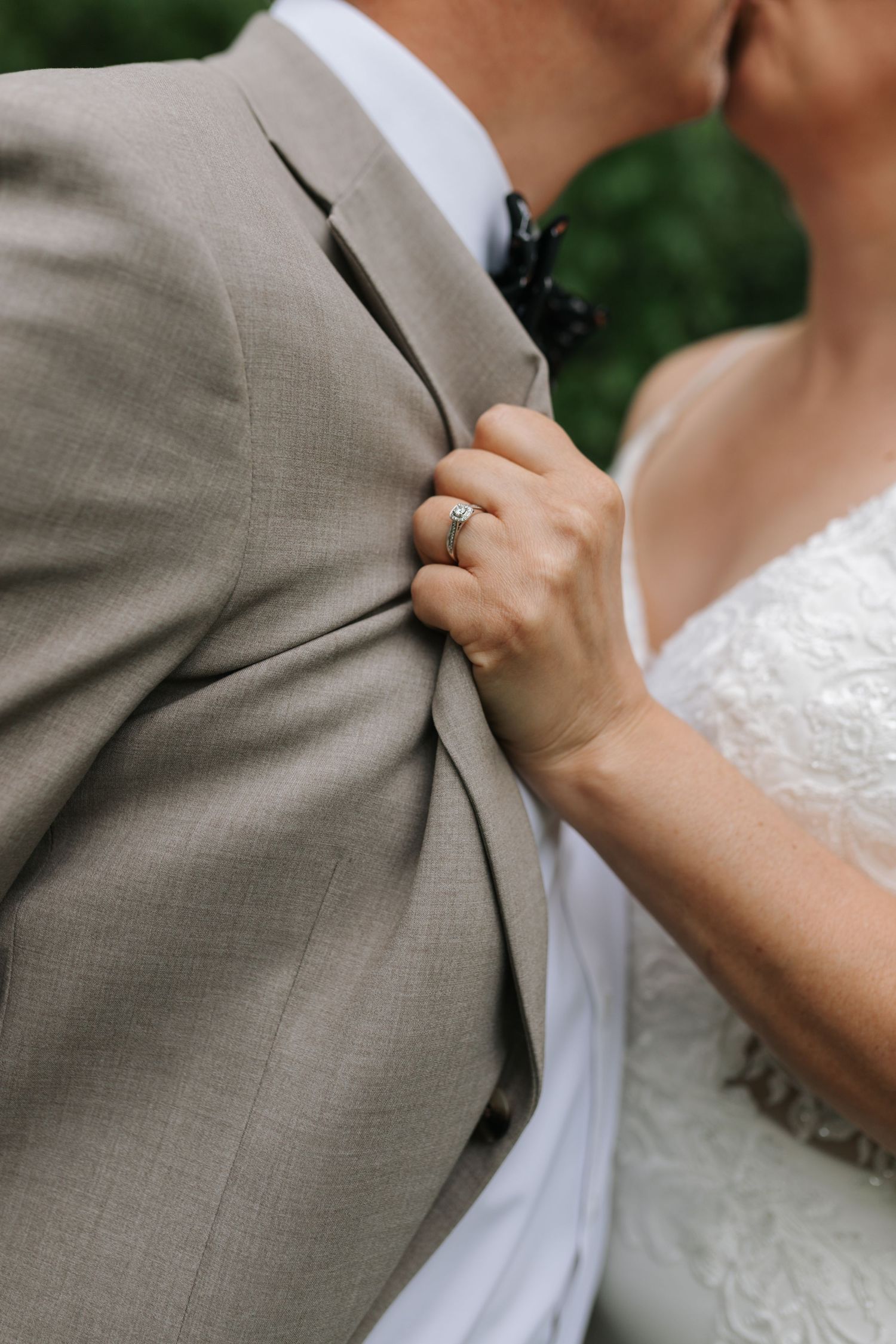 A close-up of a bride gripping her grooms tan suit jacket, showcasing her diamond engagement ring during their wedding.