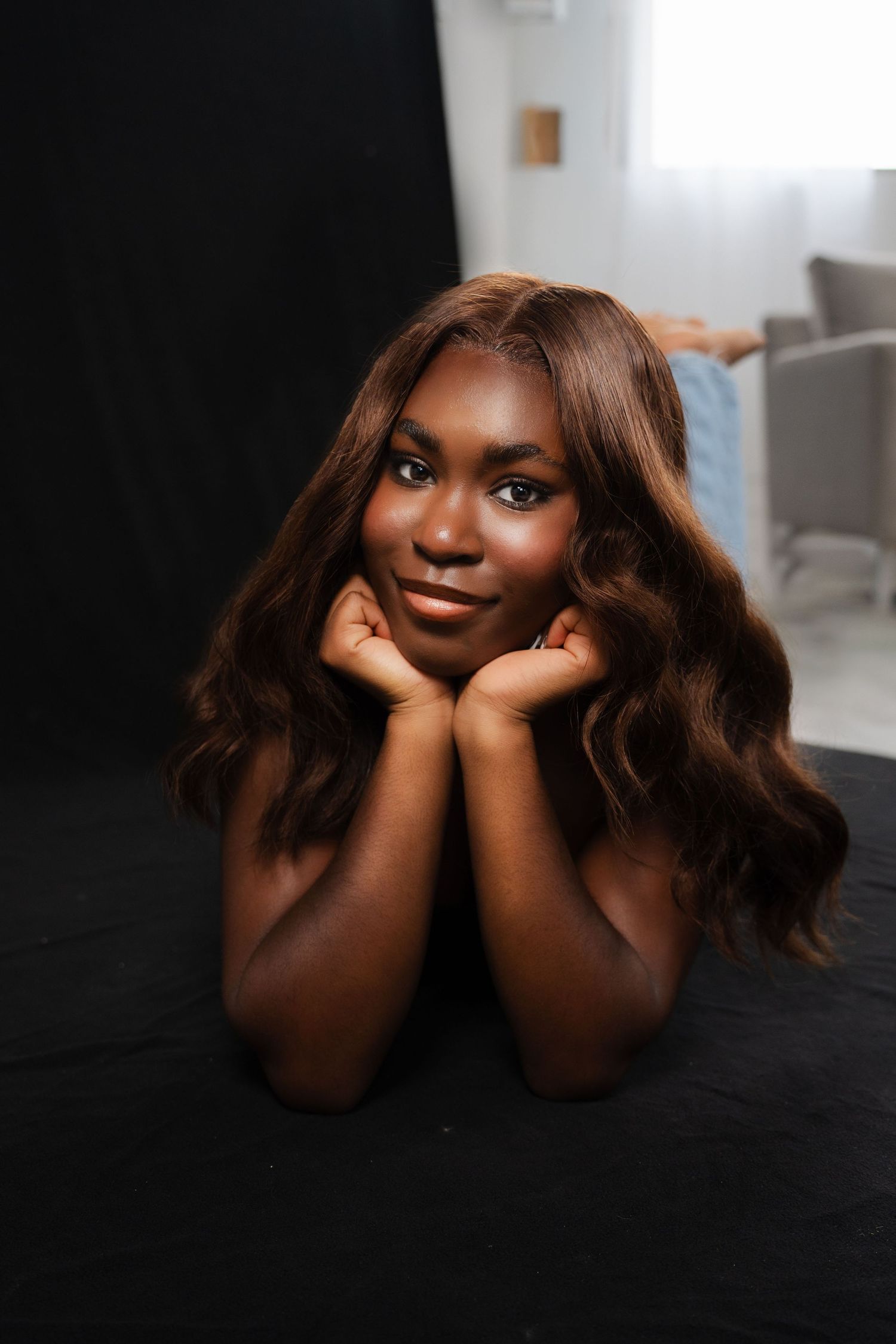 A young Black woman with long wavy brown hair rests her chin on her hands, smiling softly at the camera.