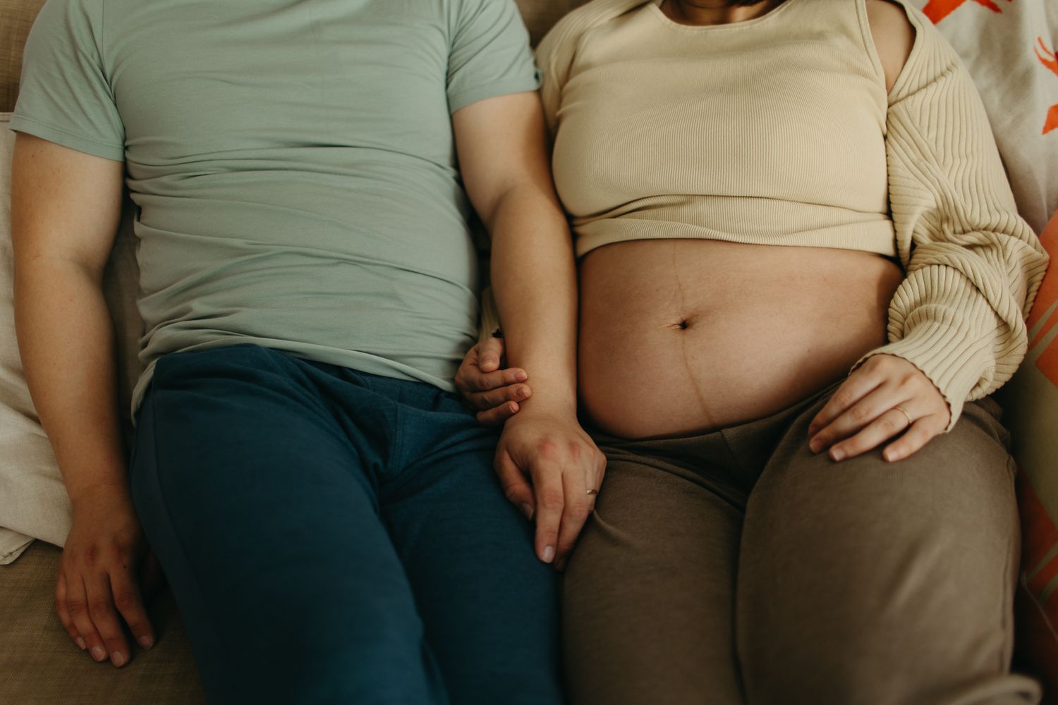 Pregnant woman holding hands with partner while relaxing on a couch, showing baby bump in a cozy setting.